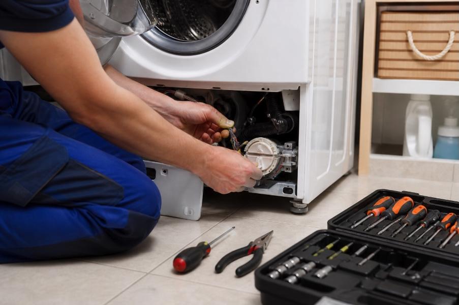 Close-up of a technician’s hands repairing the internal components of a front-loading washing machine in a clean, well-lit laundry room, with a neatly arranged set of tools on the floor nearby.